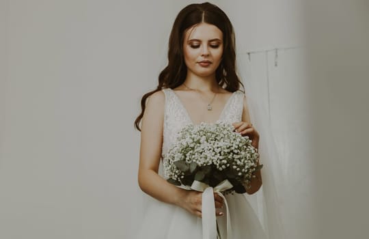 Portrait of one beautiful young Caucasian brunette bride with a white bouquet of flowers and boutonnieres in her hands, standing in the room behind the door and looking thoughtfully down, side view, close-up.