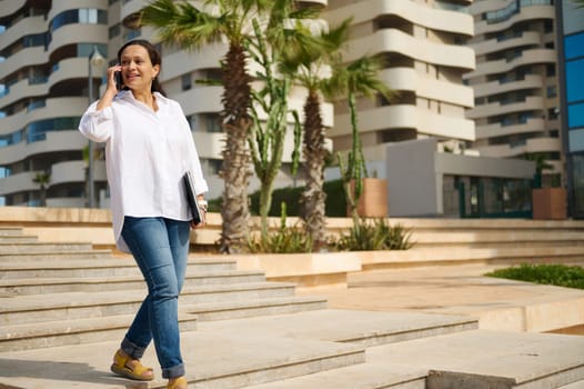 Confident young female freelance worker, business woman talks on smart mobile phone while going to the office, holding laptop against cityscape background. People. Recruitment. Employment and career