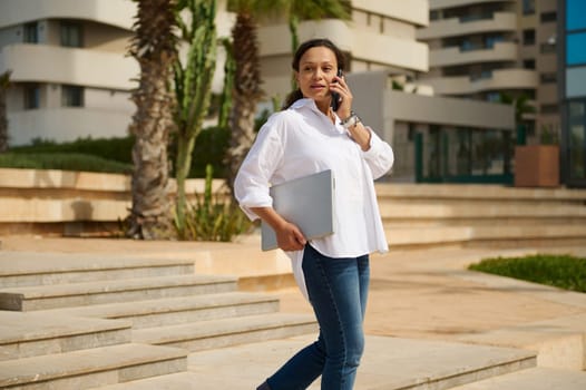 Portrait of charming young adult woman dressed in stylish white shirt and blue jeans, holding laptop and looking confidently into the distance while walking down the steps against cityscape background