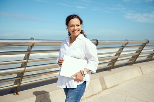 Attractive Middle Eastern pretty woman, confident successful freelancer, manager enjoying beautiful sunny day, strolling the seashore promenade with laptop in her hands, cutely smiling looking aside