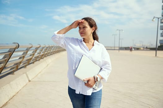 Confident relaxed young multi ethnic woman freelancer in casual clothes, holding laptop and looking into the distance, standing on the promenade against seashore background. Copy advertising space