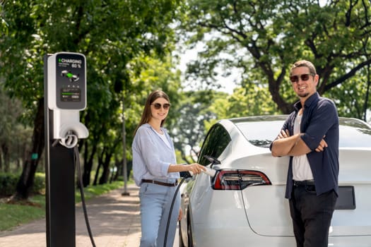 Lovely young couple wearing sun glasses recharging battery for electric car during road trip travel EV car in natural forest or national park. Eco friendly travel during vacation and holiday. Exalt