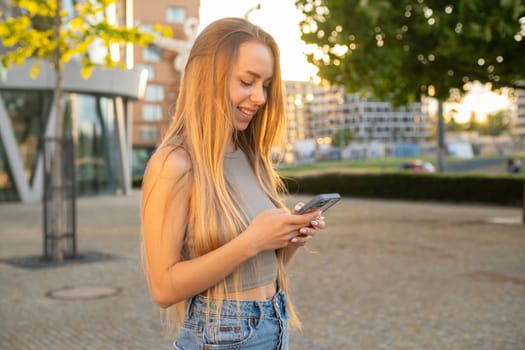 Cheerful young woman widely smiles at phone screen texting message to friend. Laughing lady relaxes in peaceful park after work surfing internet