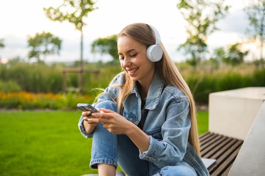 Joyful lady in headphones relaxes hugging legs on park bench in summer. Young woman listens to music messaging friends at evening twilight