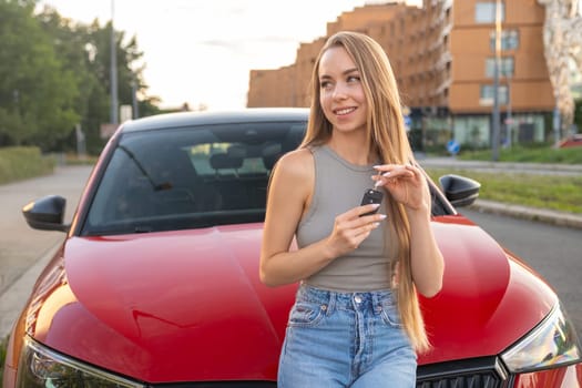 A young woman driver with blonde long hair stands before her red car, holding the key.