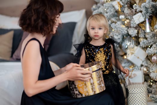 Mother daughter 2 years old Christmas tree. Both are dressed in black dresses, the mother holds the girl in her arms and both look at each other. The family celebrates Christmas