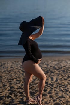 Pregnant woman in a large straw hat and black swimsuit posing on the beach