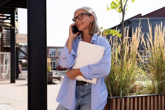 pretty fashionable middle-aged woman with gray hair dressed in summer solves business affairs with a laptop while walking on the street.