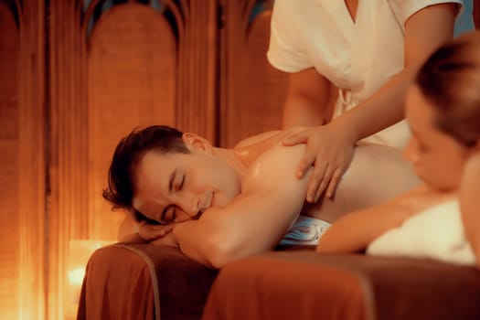 Caucasian couple customer enjoying relaxing anti-stress spa massage and pampering with beauty skin recreation leisure in warm candle lighting ambient salon spa at luxury resort or hotel. Quiescent