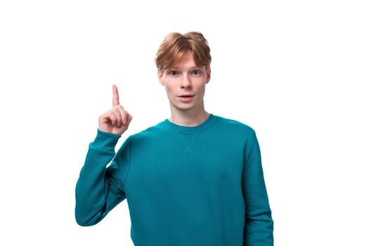 young caucasian student guy with red hair dressed in a blue sweater raised his thumb up on a white background with copy space.