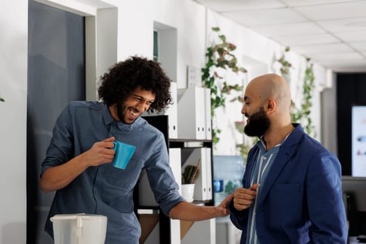Corporate colleagues telling joke and laughing while drinking coffee in coworking space. Smiling young arab start up company employees enjoying work break in business office