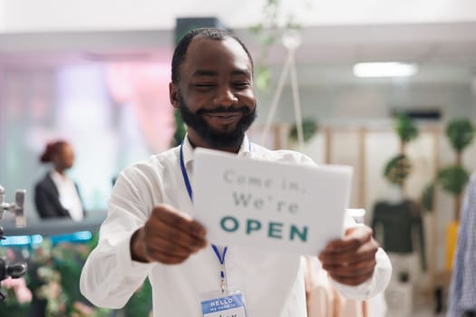 Clothes store smiling cheerful assistant hanging opening sign on front door. Happy african american fashion boutique manager holding sign board with come in text message