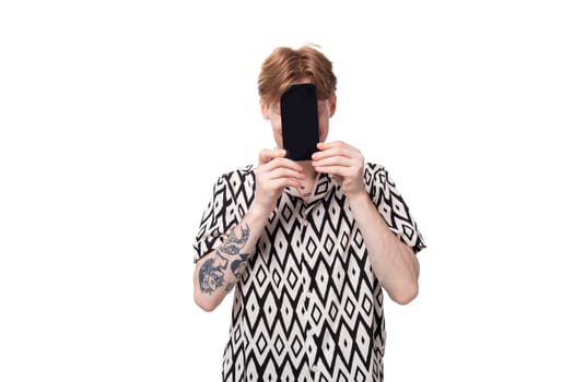 young smiling red-haired man dressed in a summer short-sleeved shirt in black and white holds a smartphone with a mockup.
