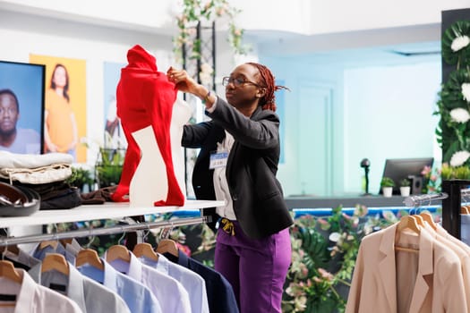 Clothes retail store assistant wearing casual red blouse on mannequin to showcase outfit for customers. Fashion boutuque african american woman employee dressing dummy model