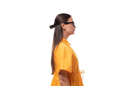 European young woman with black hair dressed in an orange summer dress stands sideways.