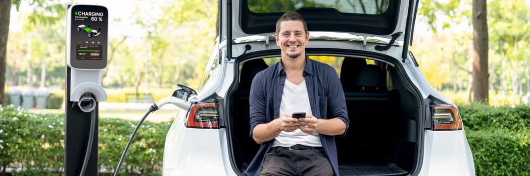 Man using smartphone online banking application to pay for electric car battery charging from EV charging station during vacation road trip at national park or summer forest. Panorama Exalt