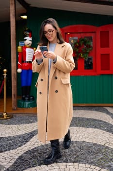 Woman in glasses using mobile phone at day in the city at Christmas holiday. Happy woman using smartphone dressed stylish trench coat looking device screen. Vertical photo full length