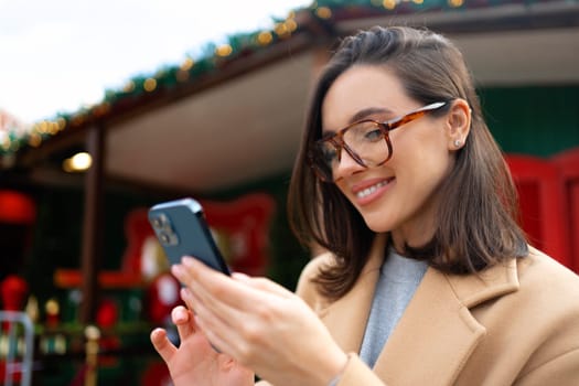 Woman in glasses using mobile phone at day in the city. Happy woman using smartphone dressed stylish trench coat looking device screen