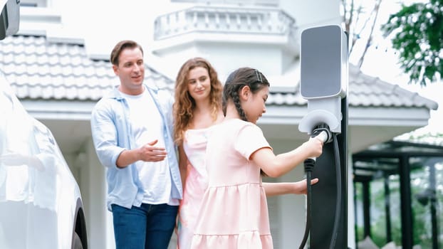 Happy little young girl learn about eco-friendly and energy sustainability as she help her family recharge electric vehicle from home EV charging station. EV car and modern family concept. Synchronos