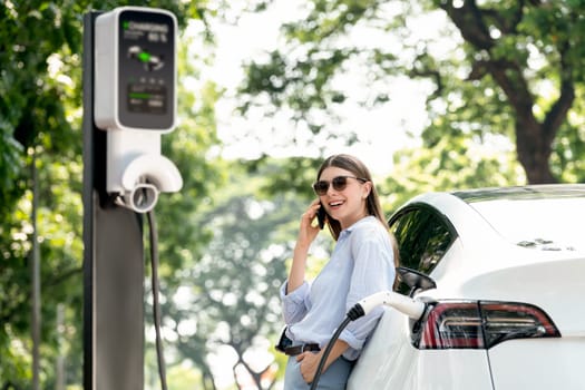 Young woman using smartphone online banking application to pay for electric car battery charging from EV charging station during vacation holiday road trip at national park or summer forest. Exalt