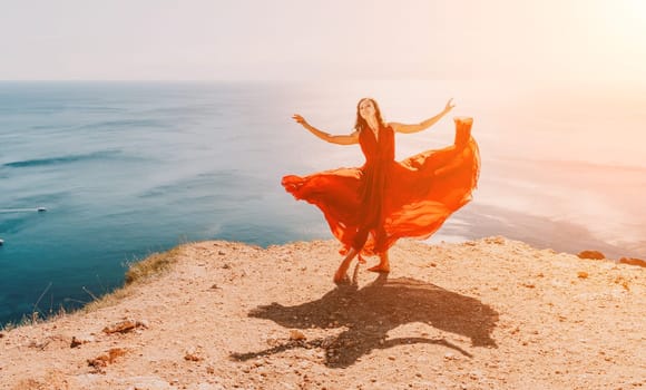 Side view a Young beautiful sensual woman in a red long dress posing on a rock high above the sea during sunrise. Girl on the nature on blue sky background. Fashion photo.