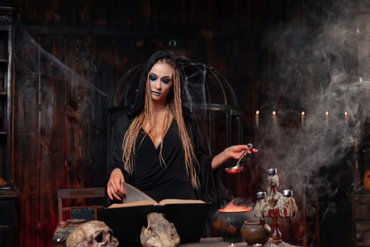 Halloween, witch use magic book and cauldron prepare poison or love potion. Black magic occult female wizard in dark room cage spider web human skull. Dressed black cloak with dreadlocks