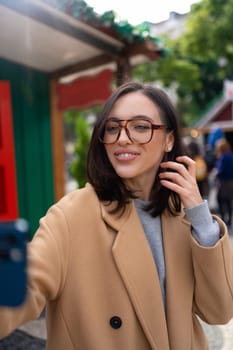 Portrait of positive woman 30s in glasses laughing while taking selfie photo on smartphone dressed stylish trench coat standing outside on autumn city street. Vertical photo
