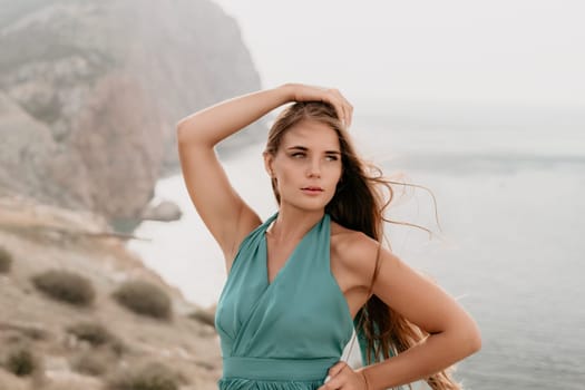 Side view a Young beautiful sensual woman in a mint long dress posing on a volcanic rock high above the sea during sunset. Girl on the nature on overcast sky background. Fashion photo