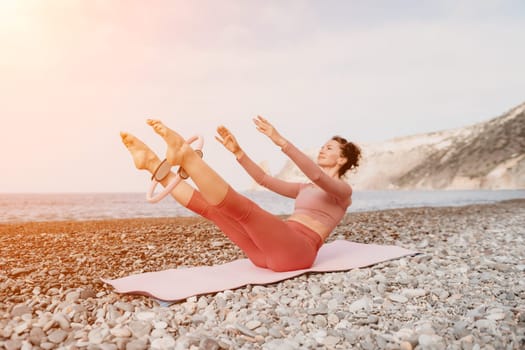 Middle aged well looking woman with black hair doing Pilates with the ring on the yoga mat near the sea on the pebble beach. Female fitness yoga concept. Healthy lifestyle, harmony and meditation.