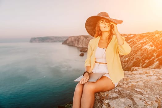 Successful business woman in yellow hat working on laptop by the sea. Pretty lady typing on computer at summer day outdoors. Freelance, travel and holidays concept.