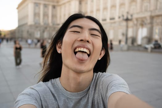 Asian young guy showing his tongue while taking selfie photo in Madrid city.
