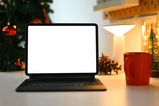 Blank display screen digital tablet and cup of hot chocolate on white table in decorated living room for Christmas.