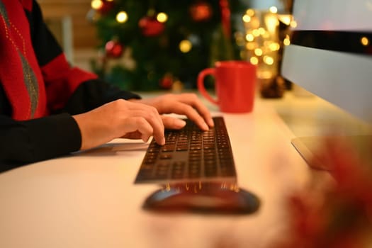 Closeup woman hands typing on computer keyboard against Christmas tree with lights on background.