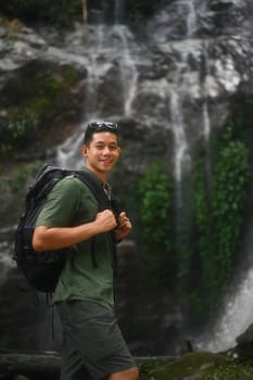 Portrait of handsome male tourist with backpack standing in front of the tropical waterfall.