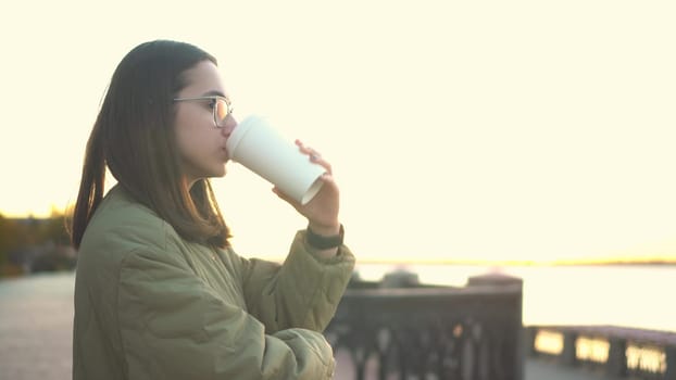 Young woman with a glass of coffee in her hand. A girl in glasses near the fence on the embankment with a hot drink in a glass. 4k