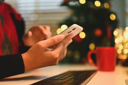 Cropped shot of young woman using mobile phone near Christmas tree at cozy home.