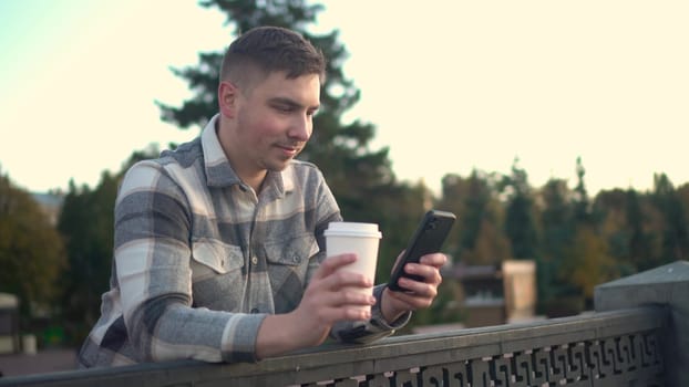 A young man is chatting on a smartphone with a glass of coffee in his hand. A man with a phone near the fence on the embankment with a hot drink in a glass. 4k