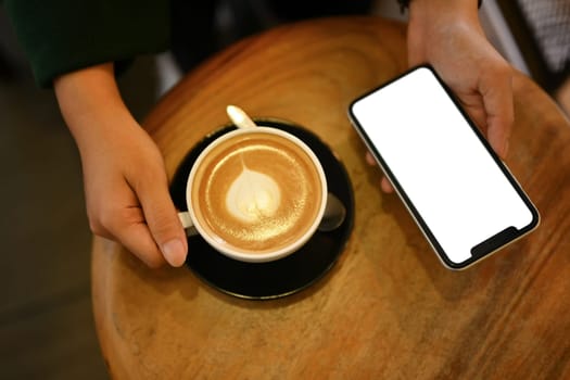 Above view of young woman holding cup of hot latte coffee and using mobile phone.