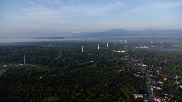 Aerial view of High voltage grid tower with wire cable at tree forest with fog in early morning. Colorful landscape with woods in fog, sunbeams, sky, forest in winter morning.