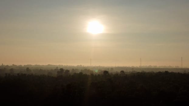 Aerial view of High voltage grid tower with wire cable at tree forest with fog in early morning. Colorful landscape with woods in fog, sunbeams, sky, forest in winter morning.