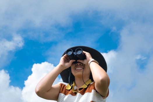 Young woman with binoculars on the mountain on a sunny day. Woman using binoculars when going hiking. Hiking woman uses binoculars to travel and has a happy smile.