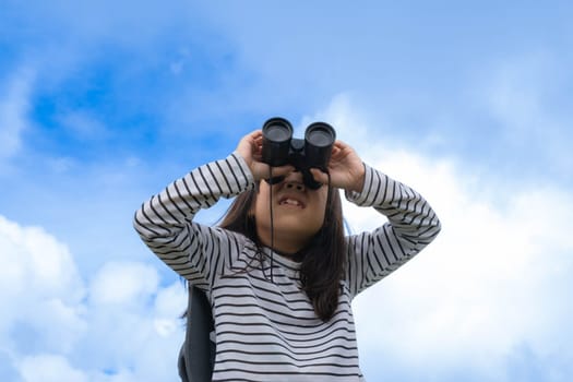 Cute little girl with binoculars on the mountain on a sunny day. Young girl uses binoculars when going hiking. Active young girl uses binoculars on a trip and smiles happily.