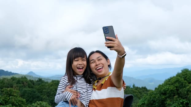 Cute little Asian girl and mother take selfie together with beautiful view of green mountains in summer. A girl traveling with mother. A woman is photographed with her daughter, Camping in the mountains with children.