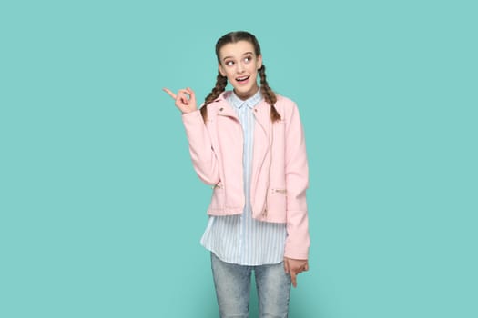 Portrait of excited smiling positive teenager girl with braids wearing pink jacket pointing index finger away, showing copy space for advertisement. Indoor studio shot isolated on green background.