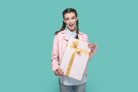 Portrait of excited positive teenager girl wearing pink jacket standing wrapped present box in hands, looking at camera, waiting holiday to give gift. Indoor studio shot isolated on green background.