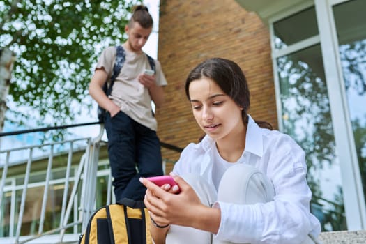 Teenage students with smartphones on the steps of educational building, in focus teenager girl high school age using gadget. Adolescence, youth, education, lifestyle, technology concept