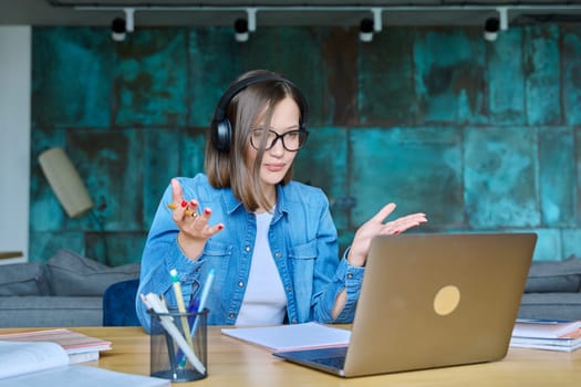 Female university student with headphones having online lesson lecture, video conference, sitting at table at home, looking laptop screen. Internet technologies in education e-learning virtual classes