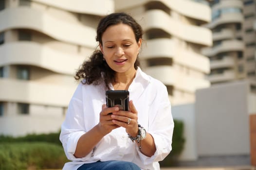 Portrait of confident casual woman using smartphone, checking mobile application, scrolling news feed, checking social media content, smiling, sitting outdoors against modern buildings backdrop