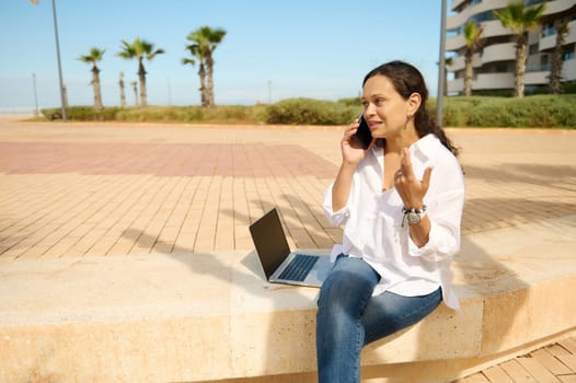 Confident young woman in casual white shirt and blue jeans, talking on mobile phone while working online outdoors, negotiating new startup with business partners. People. Remote job. Freelance work