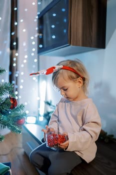 Little girl in a hoop examines red garlands-beads in a box while sitting on a low locker near the Christmas tree. High quality photo